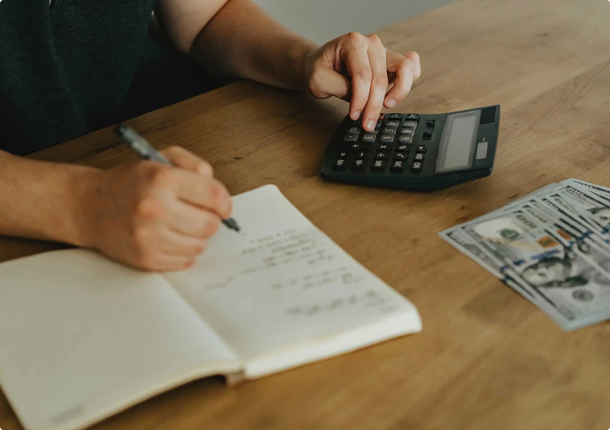 Hand writing in a notebook with a pen, a calculator, and U.S. dollar bills, representing financial record-keeping and accounting services.