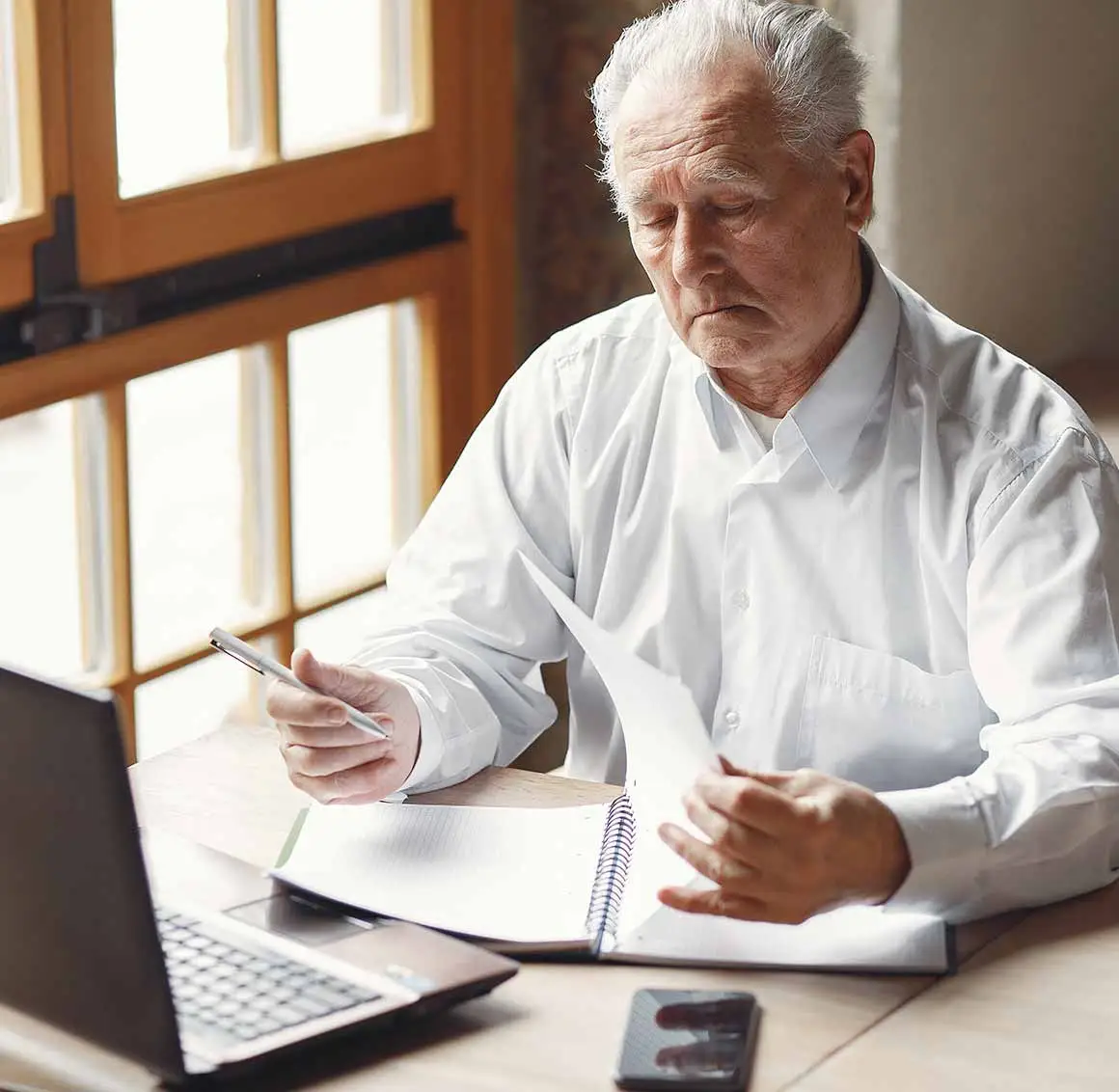 Senior man wearing a shirt, sitting at a table with an open laptop and financial documents, looking concerned while reviewing paperwork.