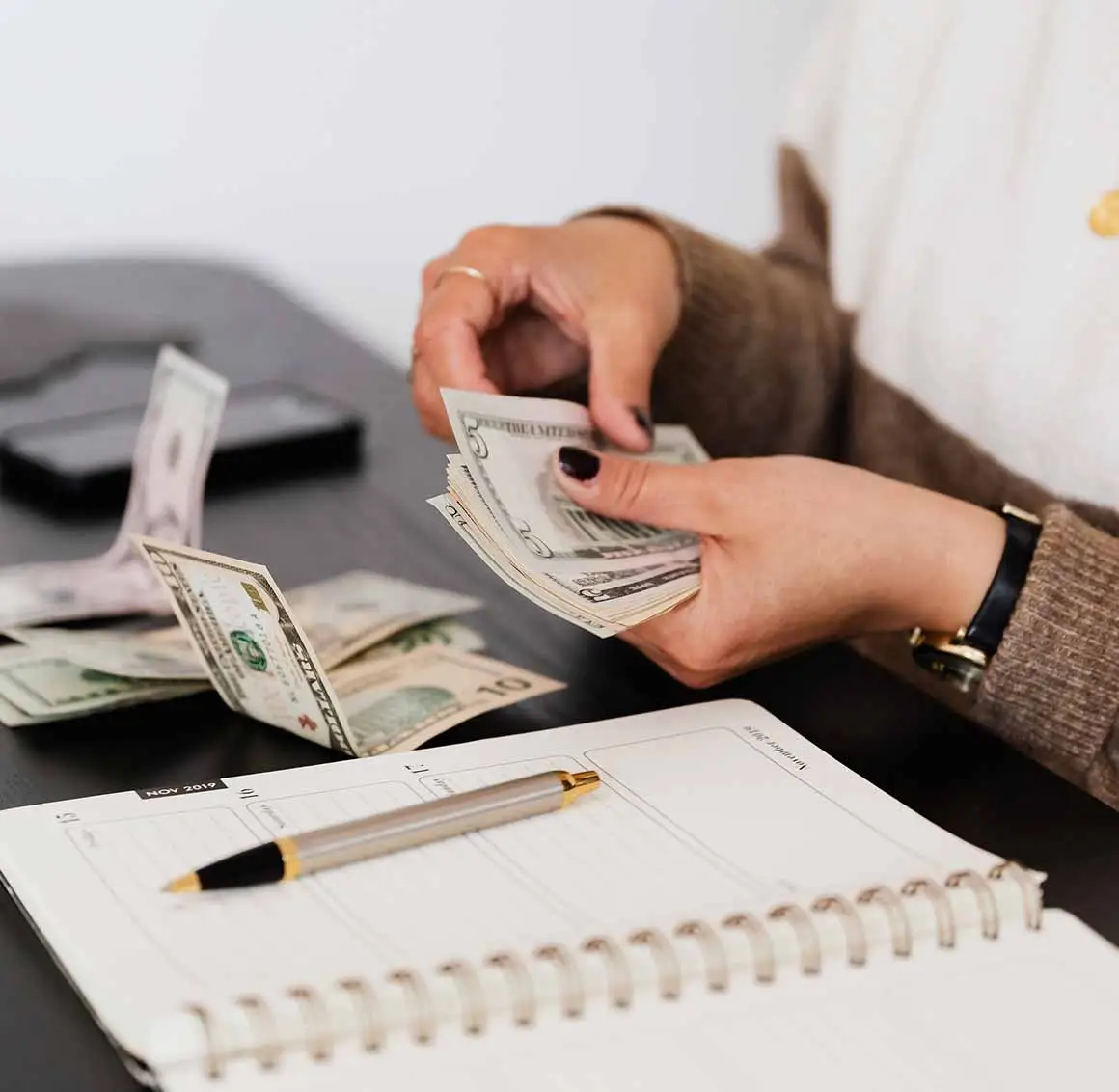 Female hands wearing a sweater and watch, counting U.S. dollar bills on a desk with additional cash, a pen, and a notebook, symbolizing payroll management.